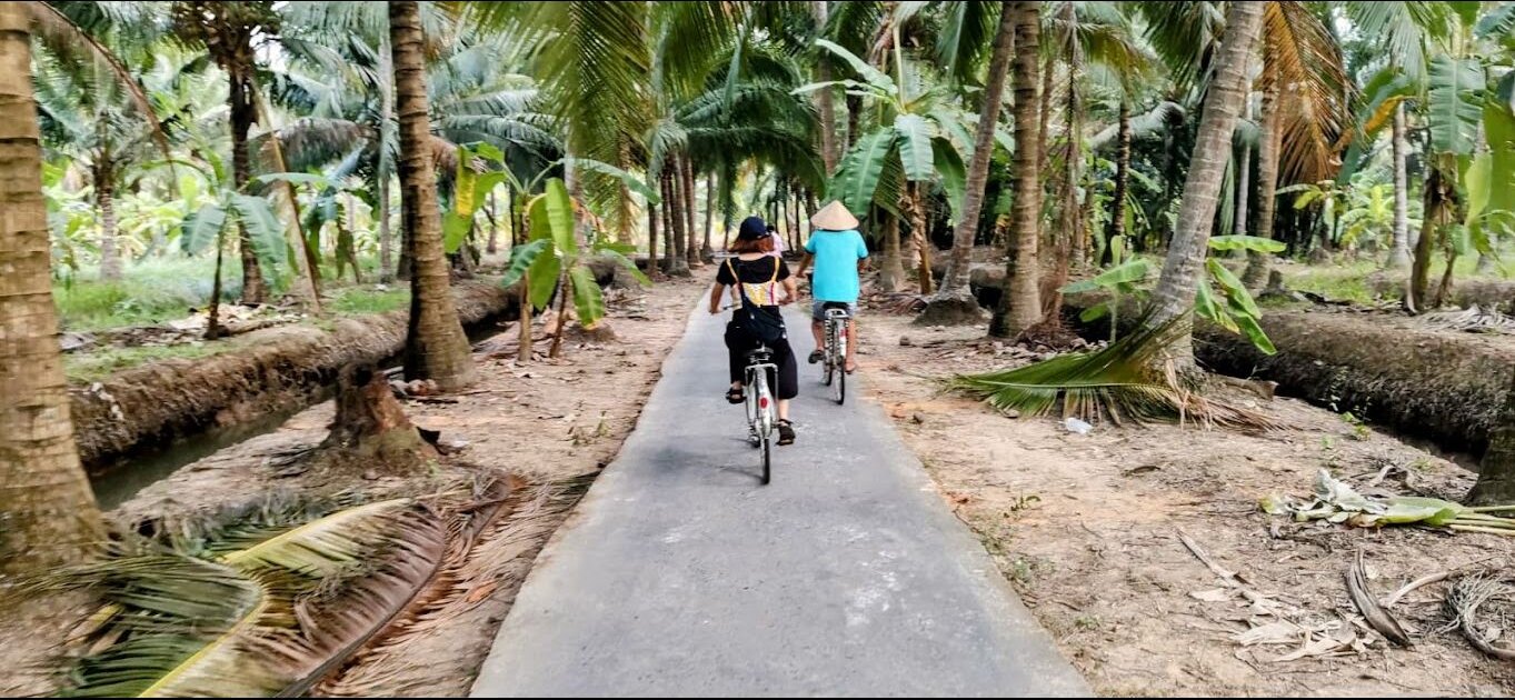 Two travelers cycling along a narrow lane through coconut gardens